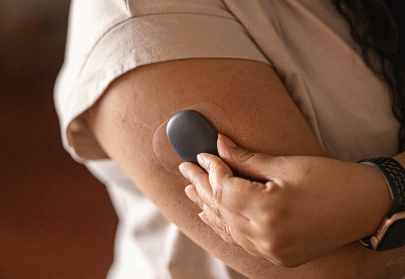 Hands attach a small, round device to an upper arm inside a short-sleeved shirt, against a blurred background.