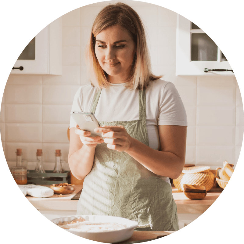 Eversense CGM user in an apron uses a smartphone in a bright kitchen, with baking ingredients on the counter.