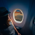 Woman in hat gazes out airplane window; exterior shows runway and sunset, creating a serene travel ambiance.